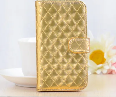 A gold quilted phone case is displayed upright on a light-colored surface, with a white teacup and saucer to its left and blurred flowers to its right.