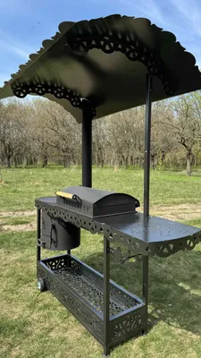 A black metal grill station with a roof and a charcoal grill is set up outdoors on a grassy area with trees in the background.