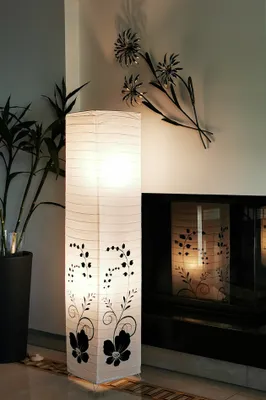 A tall, white paper lantern with black floral designs stands in a room next to a fireplace and a plant.