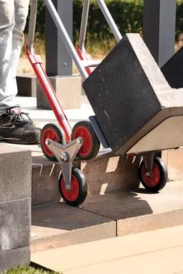 A person is using a sack truck with a large stone block on it to go up some steps.