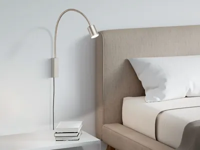 A bedside lamp is mounted on the wall above a nightstand with books, next to a bed with a headboard and pillows.