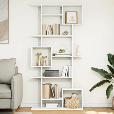 A white, modern, modular bookshelf filled with books and decorative items, placed in a living room setting next to a sofa and a large plant.