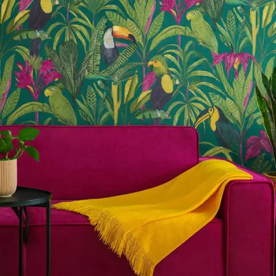 A bright yellow throw blanket is draped over a plush magenta sofa, set against a vibrant tropical wallpaper featuring parrots and toucans.
