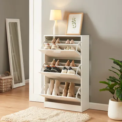A white shoe cabinet with multiple compartments filled with shoes, placed in a hallway next to a mirror and a plant.