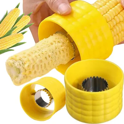A hand is using a yellow corn peeler to remove kernels from a cob of corn. Other parts of the peeler and some corn cobs are visible in the foreground and background.