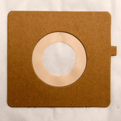 A square brown paper vacuum cleaner bag with a circular opening in the center, lined with a beige ring and covered by a white filter material. The bag is lying on a white textured surface.