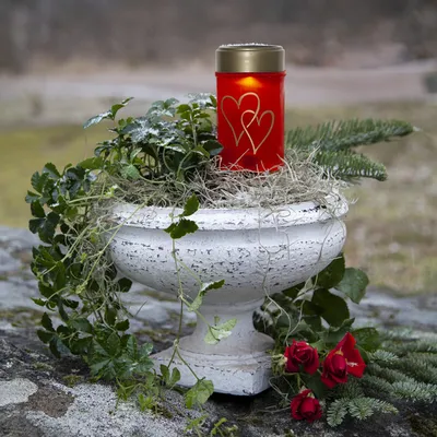 A red candle with gold hearts sits in a white urn filled with greenery and straw, placed on a stone surface.