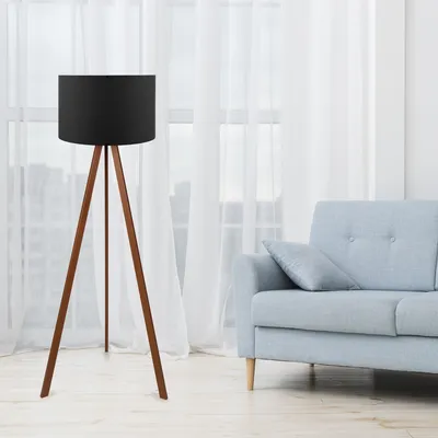 A floor lamp with a black shade and wooden tripod base stands in a living room next to a light blue sofa.