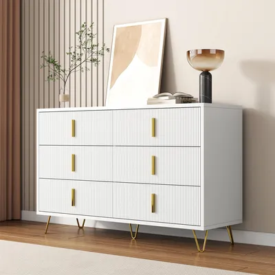 A white dresser with gold handles and legs, decorated with a framed picture and a decorative bowl.
