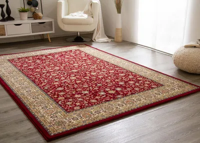 A red oriental rug with a floral pattern and a beige border is placed on a wooden floor in a living room. A white armchair and a cabinet are visible in the background.