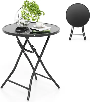 A round, black, folding bistro table with a glass top, set against a white background. A small potted plant, a cup, and some books are on the table. A second, folded table is shown in the background.