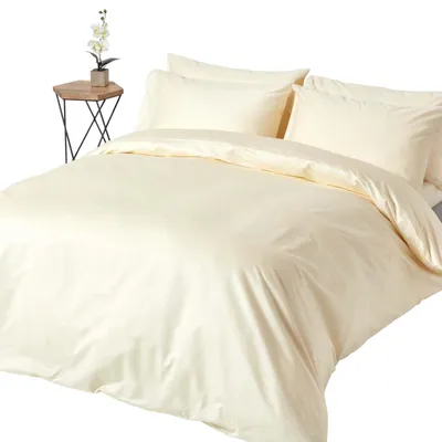 A bed with cream-colored bedding, including a duvet and pillows, on a white background. A small side table with a plant is visible to the left.