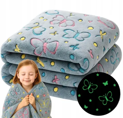 A young girl is wrapped in a soft, gray blanket with a butterfly pattern. The blanket is folded in the background, and a circular inset shows the blanket's glow-in-the-dark feature.