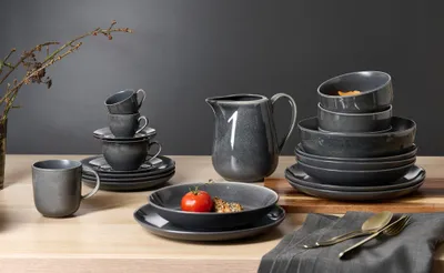 A collection of dark gray ceramic tableware, including plates, bowls, cups, and a pitcher, arranged on a wooden surface.