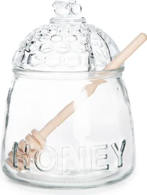 A clear glass honey jar with a honeycomb lid and a wooden honey dipper inside.