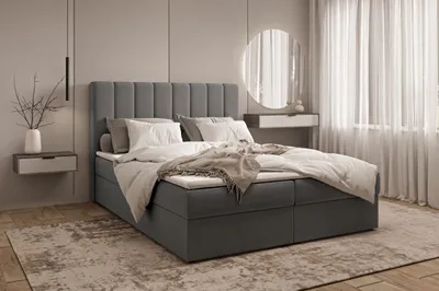 A bedroom scene featuring a large bed with a padded headboard, dressed in white linens and a beige throw blanket. A bedside table with a vase and branches is visible to the left, and sheer curtains are in the background to the right.