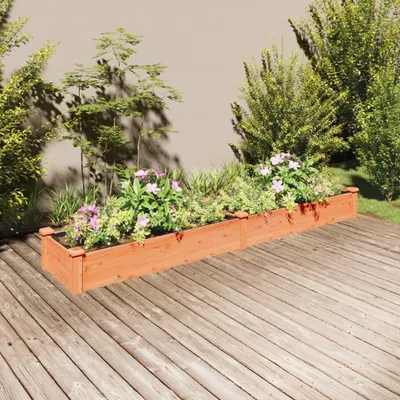 A wooden planter box filled with plants and flowers sits on a wooden deck, with a wall and bushes in the background.