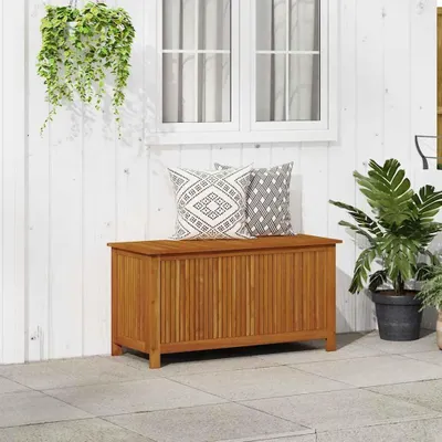 A wooden storage box with two decorative pillows on top, placed on a patio next to a white wall with a window and a hanging plant. A large potted plant is to the right of the box.