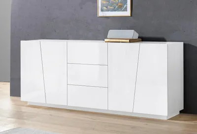 A white high-gloss sideboard with a modern, geometric design is shown in a room with a dark gray wall and a wooden floor. Decorative items are placed on top of the sideboard.