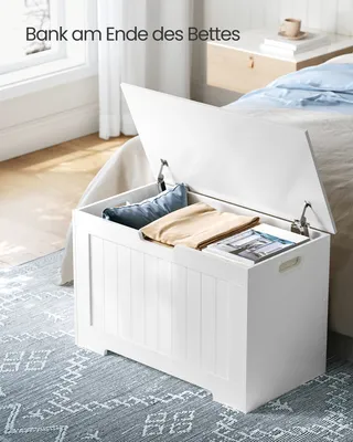 A white storage bench with its lid open, placed at the foot of a bed in a bedroom. Pillows and magazines are inside the bench.