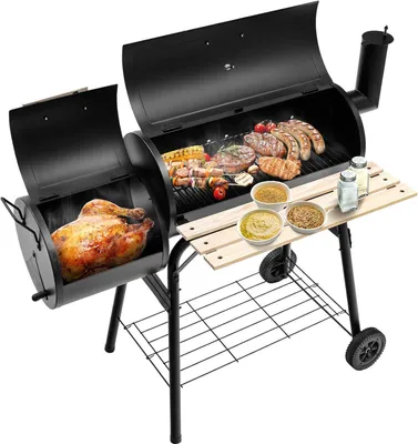 A black charcoal grill with two chambers, one containing a whole roasted chicken and the other containing various meats and skewers. It has a wooden side table and a bottom shelf, and is set against a white background.
