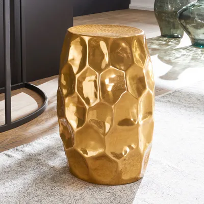 A gold, textured side table sits on a rug in a room with a dark cabinet and large glass vases in the background.