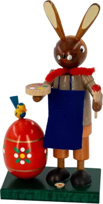Ein hölzerner Osterhase mit bemaltem Gesicht hält einen Pinsel und eine Palette in den Händen während er neben einem großen roten Osterei mit Blumenmuster steht und ein kleines gelbes Küken auf dem Ei sitzt
