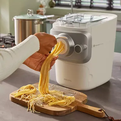 A person is using a white pasta maker to extrude fresh spaghetti onto a wooden cutting board.
