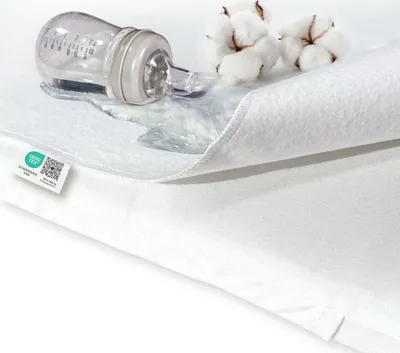 A close-up of a white waterproof mattress protector with a baby bottle and spilled liquid on it, along with cotton balls.