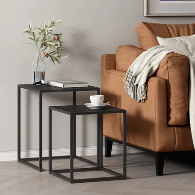 Two nesting tables with a book and a cup of coffee on them, next to a brown sofa with a blanket.