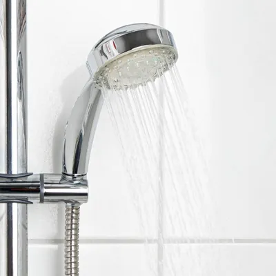 A close-up of a chrome shower head with water spraying from it, set against a white tiled wall.