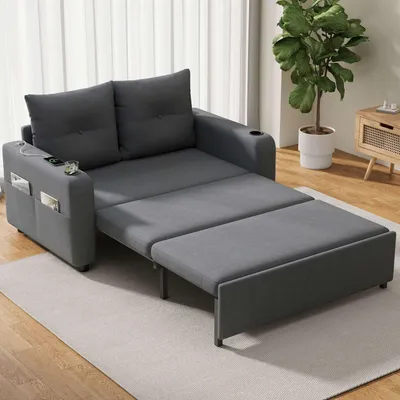 A gray sofa bed is shown in a living room setting with a plant and a side table in the background.