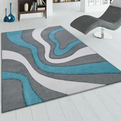 A modern rug with a wavy abstract design is displayed on a light-colored wooden floor in a room setting. A grey chaise lounge is visible in the background.