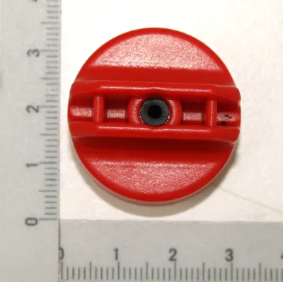 A red cap with a black center is placed on a white surface next to a ruler.