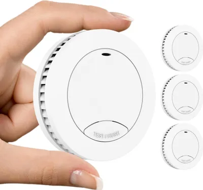 A hand is holding a white smoke detector, with three more smoke detectors visible in the background.