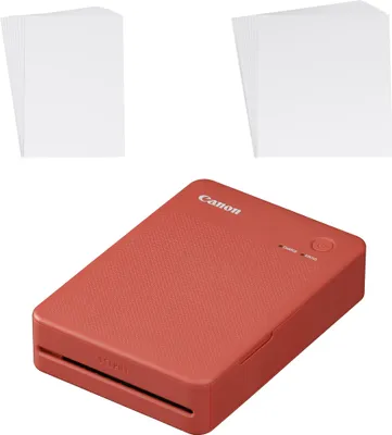 A red Canon Selphy QX20 mobile photo printer is shown with two stacks of white paper in the background.