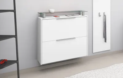 A white wall-mounted shoe cabinet with two drawers and a glass shelf on top, next to a black shelving unit.