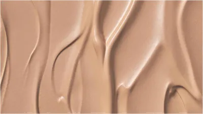 A close-up, abstract texture shot of a light beige liquid, likely foundation or BB cream, with visible swirls and highlights.