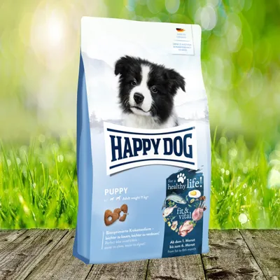 A bag of Happy Dog puppy food is shown in front of a blurred green grassy background with bokeh lights.