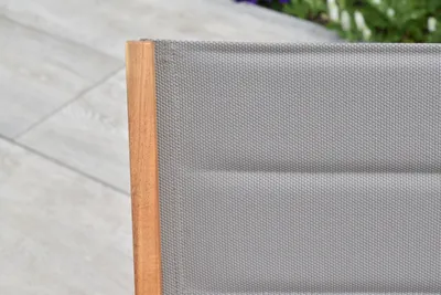 Close-up of a chair's wooden frame and grey fabric seat, with a tiled patio and greenery in the background.
