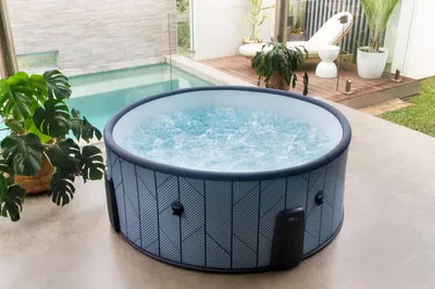 An inflatable hot tub filled with bubbling water is placed on a patio next to a swimming pool. The background shows a wooden deck with lounge chairs and potted plants.