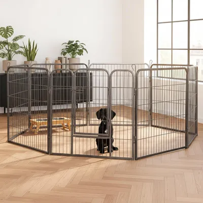A black dog sits inside a metal playpen set up in a living room with plants and furniture in the background.