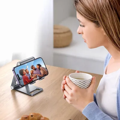 A woman holds a cup of coffee while looking at a phone on a stand.