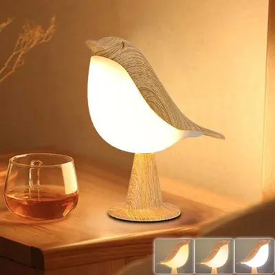 A wooden bird-shaped table lamp with a glowing white light sits on a wooden surface next to a glass of amber liquid. The background is a warm, neutral tone.