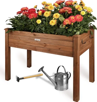 A wooden raised garden bed filled with red and yellow flowers, with a small rake and watering can placed in front of it.