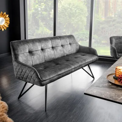 A dark gray velvet bench with a tufted back and diamond stitching on the sides, placed in a modern living room setting with large windows overlooking greenery.
