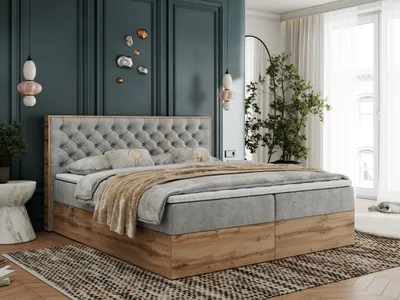 A bedroom scene featuring a large boxspring bed with a tufted headboard and wooden base, decorated with pillows and a throw blanket. The room has dark teal walls, decorative wall art, pendant lights, and a large plant near a window with sheer curtains.