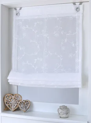 A white sheer Roman blind with embroidered floral pattern hangs in a window frame, with decorative heart-shaped objects on the windowsill below.