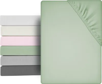 A stack of folded fitted sheets in various colors, with a single fitted sheet in light green prominently displayed in the foreground, showing its elasticized corner.