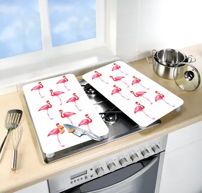 Two white stove top covers with a flamingo pattern are placed on a kitchen stove, next to a sink and a window. A pot is visible in the background.